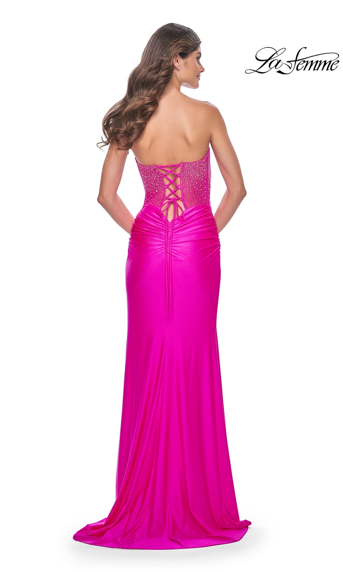 A woman stands with her back to the camera in the La Femme Strapless Hot Pink Long Prom Dress 32326, featuring a beaded corset bodice, lace-up back, and ruched detailing.
